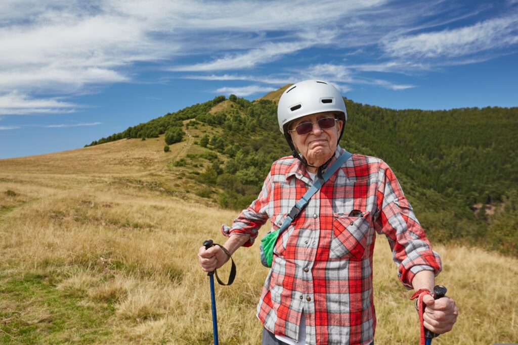 Older man wearing a helmet and sunglasses hiking with trekking poles on a grassy mountain trail